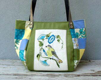 May include: A green patchwork tote bag with a floral and bird embroidery design. The bag has brown leather straps and a white background for the embroidery.