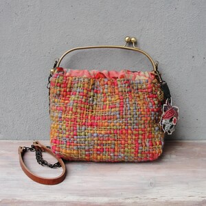K&ouml;nnte beinhalten: Eine kleine gewebte Handtasche mit einem farbenfrohen geometrischen Muster in Orange-, Rosa-, Gr&uuml;n- und Blaut&ouml;nen. Die Tasche hat einen Messingrahmen und einen Kettenriemen.