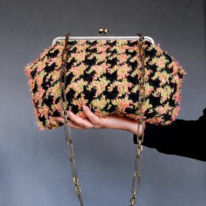 May include: A black and pink houndstooth clutch bag with a gold chain strap. The bag is held in a hand.
