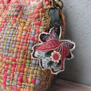 K&ouml;nnte beinhalten: Eine bunte gewebte Tasche mit einem floralen Stoffanh&auml;nger, der an einem Metallverschluss befestigt ist. Die Tasche besteht aus verschiedenen Garnen in den Farben Rosa, Orange, Gelb, Gr&uuml;n und Blau.
