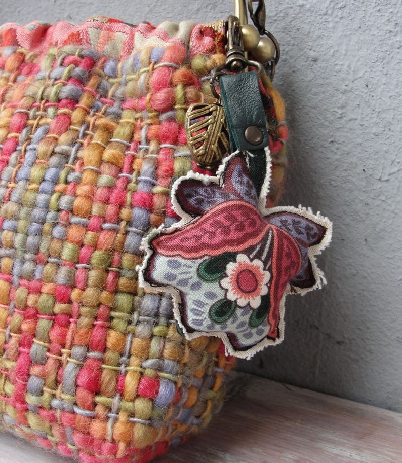 K&ouml;nnte beinhalten: Eine bunte gewebte Tasche mit einem floralen Stoffanh&auml;nger, der an einem Metallverschluss befestigt ist. Die Tasche besteht aus verschiedenen Garnen in den Farben Rosa, Orange, Gelb, Gr&uuml;n und Blau.