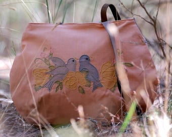 May include: A brown canvas tote bag with a strap. The bag has an embroidered design of two birds perched on a branch with yellow flowers.