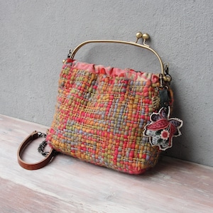 K&ouml;nnte beinhalten: Eine gewebte Wandteppich-Handtasche mit einem Messingverschluss und einem Blumenanh&auml;nger. Die Tasche ist eine Mischung aus rosa, orange, gelb und gr&uuml;nen Farben.