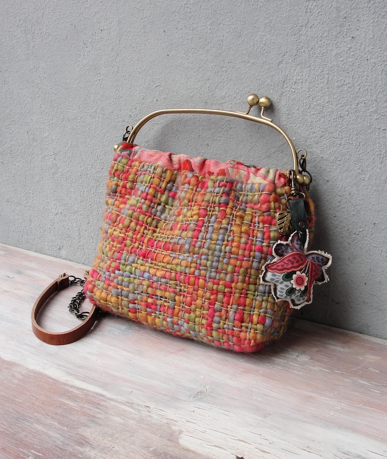 K&ouml;nnte beinhalten: Eine gewebte Wandteppich-Handtasche mit einem Messingverschluss und einem Blumenanh&auml;nger. Die Tasche ist eine Mischung aus rosa, orange, gelb und gr&uuml;nen Farben.