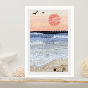 May include: A framed textile art piece depicting a coastal scene with a peach-coloured sky, a red sun, and two black birds. The ocean is rendered in layers of blue fabric and white lace, transitioning to a sandy beach made of beige fabric with small buttons.