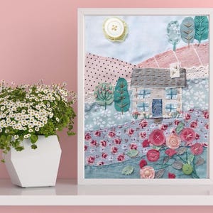 Image of Rose Cottage Slow  Stitch Kit completed in white frame. Natural fabric cottage with denim blue door pale blue windows and taupe roof. Set on fabric landscape of soft greens, floral prints, pink stripe and spot with stitched roses and trees.