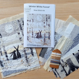 May include: A slow stitch kit titled 'Winter White Forest' featuring a forest scene with rabbits. The kit includes fabric pieces in various patterns, embroidery floss, and a printed guide. The color palette includes white, gray, and beige.