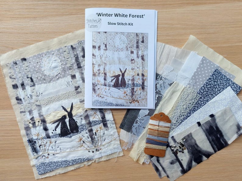 May include: A slow stitch kit titled 'Winter White Forest' featuring a forest scene with rabbits. The kit includes fabric pieces in various patterns, embroidery floss, and a printed guide. The color palette includes white, gray, and beige.