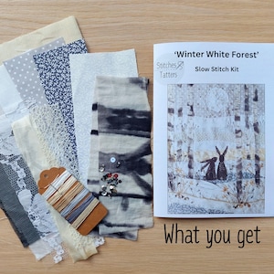 May include: A 'Winter White Forest' slow stitch kit with fabric swatches in various shades of white, gray, and blue, including lace and patterned textiles. The kit includes a booklet with a forest scene featuring rabbits and the text "What you get".