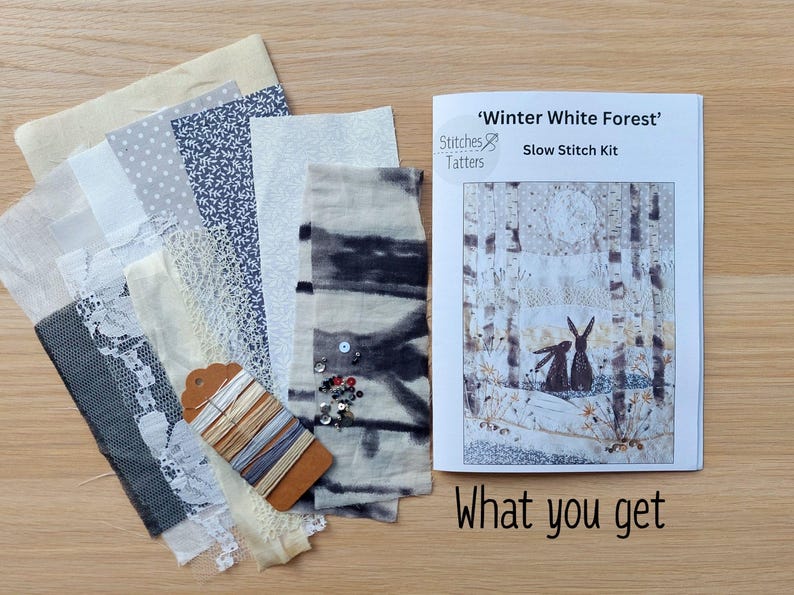 May include: A 'Winter White Forest' slow stitch kit with fabric swatches in various shades of white, gray, and blue, including lace and patterned textiles. The kit includes a booklet with a forest scene featuring rabbits and the text "What you get".