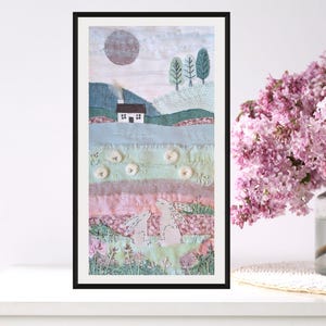 May include: Framed textile art showing a landscape with a house, trees, and rabbits. The artwork uses a soft palette of blues, greens, and pinks. The piece is framed in black and sits on a white surface. The artwork is approximately 20 cm x 40 cm.