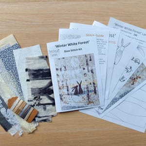 May include: A 'Winter White Forest' slow stitch kit with fabric samples, thread, and pattern guides. The kit includes various patterned fabrics in white, gray, and blue, along with a spool of thread and a tag. The pattern guides feature illustrations of rabbits in a winter forest.