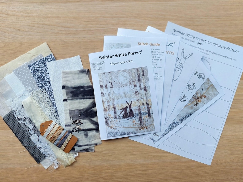 May include: A 'Winter White Forest' slow stitch kit with fabric samples, thread, and pattern guides. The kit includes various patterned fabrics in white, gray, and blue, along with a spool of thread and a tag. The pattern guides feature illustrations of rabbits in a winter forest.