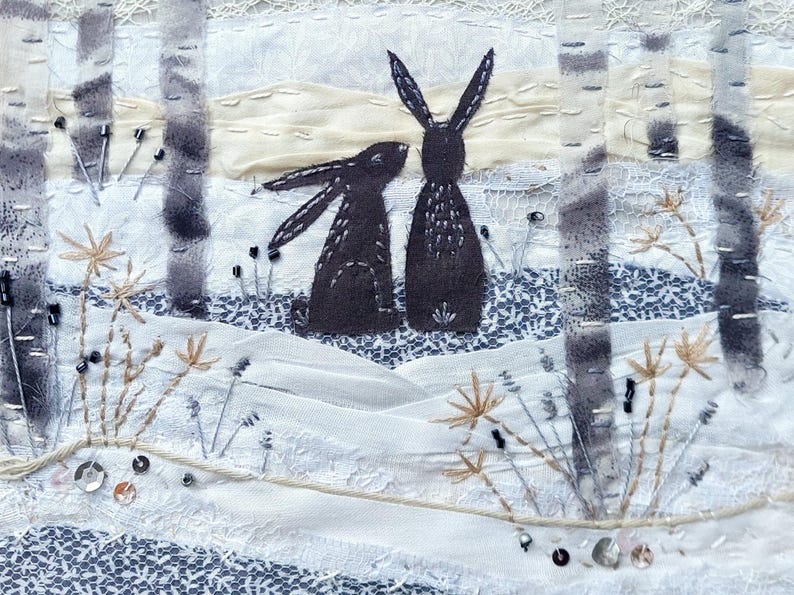 May include: Textile art piece featuring two dark gray rabbits in a winter scene. The artwork includes birch trees, embroidered details, and a snowy landscape with floral accents. The piece is predominantly white, gray, and beige.