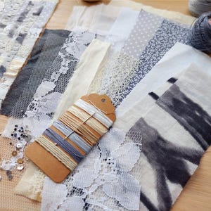May include: A collection of fabric swatches in various textures and patterns, including lace, dotted, and floral designs. A tag with assorted threads is in the center. Two balls of yarn are visible in the background. The color palette is primarily neutral with gray and cream accents.
