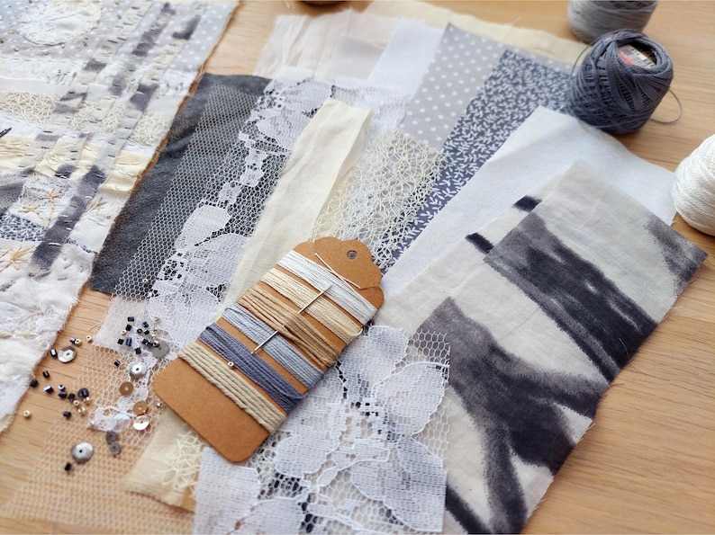 May include: A collection of fabric swatches in various textures and patterns, including lace, dotted, and floral designs. A tag with assorted threads is in the center. Two balls of yarn are visible in the background. The color palette is primarily neutral with gray and cream accents.