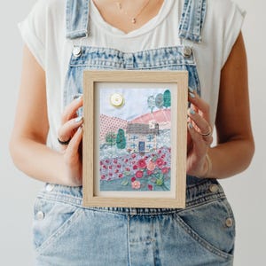 May include: A framed fabric art piece depicting a small blue house with a white door and windows, surrounded by pink and red flowers and green trees. The house is set against a backdrop of a blue sky with a yellow sun.