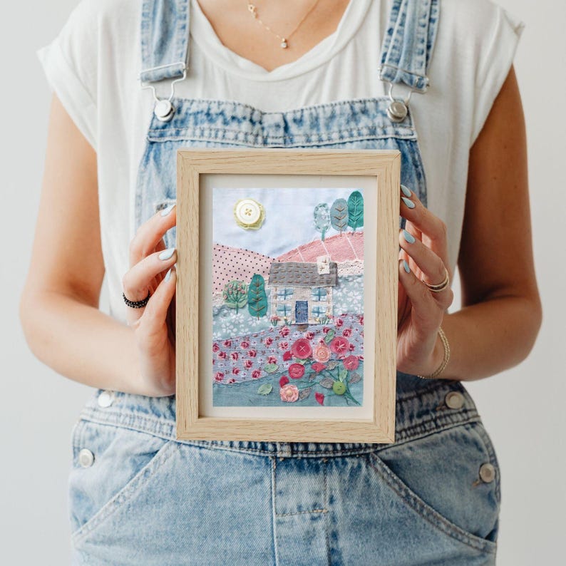 May include: A framed fabric art piece depicting a small blue house with a white door and windows, surrounded by pink and red flowers and green trees. The house is set against a backdrop of a blue sky with a yellow sun.