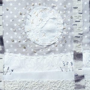 May include: Textile art piece depicting a winter scene. A white lace moon is centered above a snowy landscape with embroidered birch trees. The background is a light gray fabric with white polka dots. Delicate embroidery and lace details.