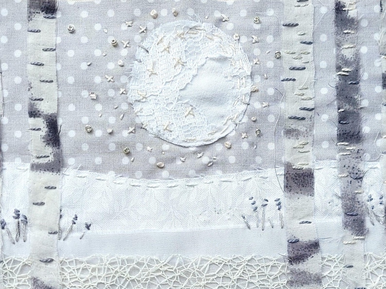 May include: Textile art piece depicting a winter scene. A white lace moon is centered above a snowy landscape with embroidered birch trees. The background is a light gray fabric with white polka dots. Delicate embroidery and lace details.
