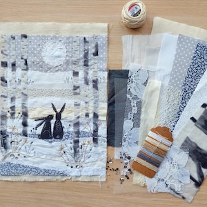 May include: Textile art piece depicting two black rabbits in a snowy forest scene with a full moon. Various fabric swatches in white, gray, and blue, along with spools of thread and a tag with thread samples, are arranged on a wooden surface.
