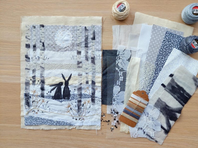 May include: Textile art piece depicting two black rabbits in a snowy forest scene with a full moon. Various fabric swatches in white, gray, and blue, along with spools of thread and a tag with thread samples, are arranged on a wooden surface.