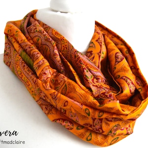 May include: A bright orange infinity scarf with a paisley pattern in shades of green, pink, and brown. The scarf is made of silk and is soft and comfortable to wear.
