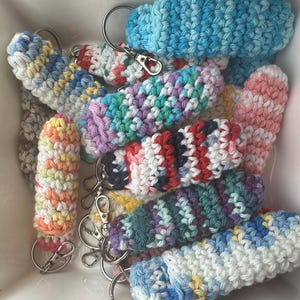 May include: A collection of colorful crocheted keychains, each with a different pattern and color combination. The keychains are made with a variety of yarn colors, including blue, pink, green, red, and yellow. The keychains are all attached to silver key rings.