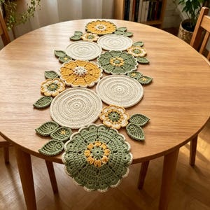 Pode incluir: Um caminho de mesa em crochê com uma série de tapetes circulares na cor creme, intercalados com vários motivos florais em tons de verde sálvia, amarelo mostarda e branco, acentuados por pequenas folhas de crochê em verde sálvia.