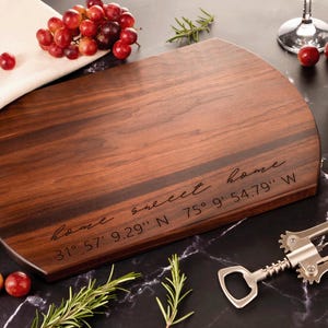 May include: A wooden cutting board with a rounded edge and a dark brown wood grain. The board is engraved with the text "home sweet home" and coordinates: "31° 57' 9.29" N 75° 9' 54.79" W".