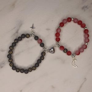 May include: Two matching beaded bracelets. One is black with gold accents and a silver cross charm. The other is red and pink with a silver crescent moon charm. Both bracelets connect with a magnetic heart.