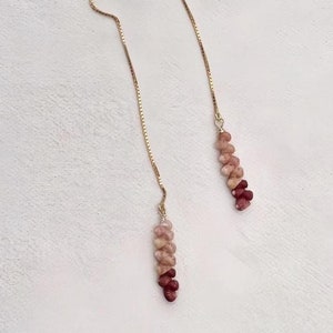 May include: A pair of gold-colored threader earrings. Each earring features a delicate chain with a cluster of pink and red gemstones at the end. The earrings are displayed on a white surface.
