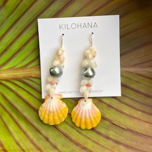 May include: Handcrafted jewelry earrings featuring yellow and pink seashell pendants. The earrings have a strand of small white shells and a gray pearl. The earrings are hanging from a white card with the text "KILOHANA" and "HANDCRAFTED JEWELRY".