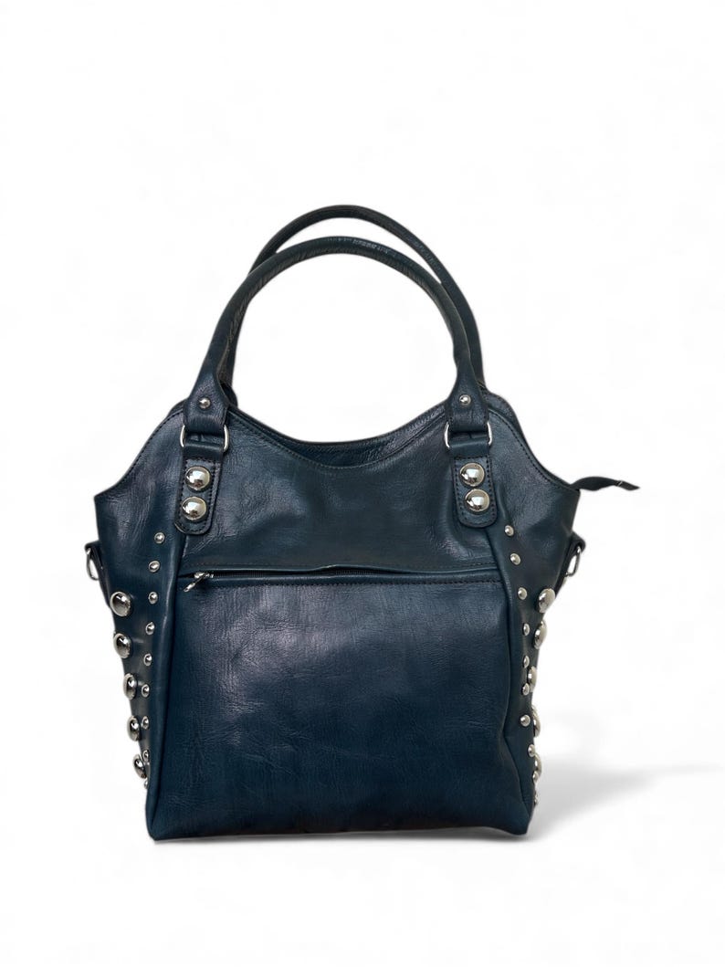 May include: A dark blue leather handbag with silver-colored stud accents. The bag has two top handles and a front zippered pocket. The bag is a tote style, with a structured shape and a slight curve at the top.