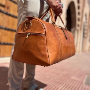 May include: A large, tan leather duffel bag with a zipper pocket and two handles. The bag is held by a person. The bag has a gold-colored zipper and hardware. The bag is made of a smooth, textured leather.