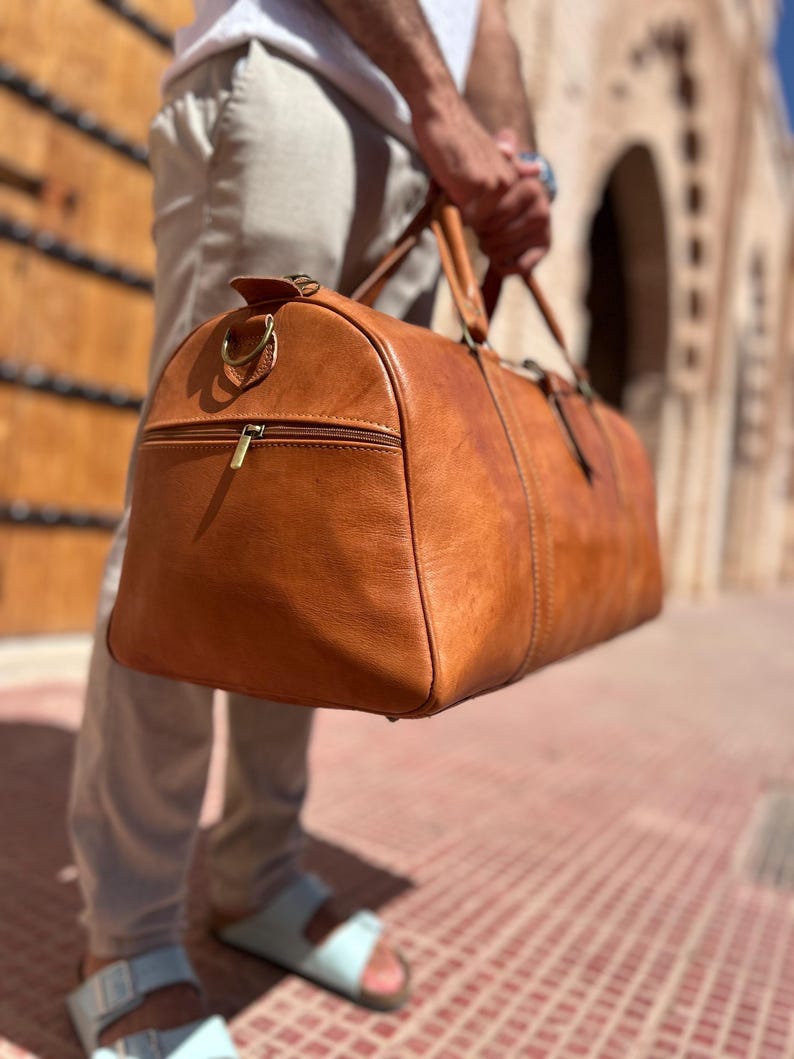 May include: A large, tan leather duffel bag with a zipper pocket and two handles. The bag is held by a person. The bag has a gold-colored zipper and hardware. The bag is made of a smooth, textured leather.