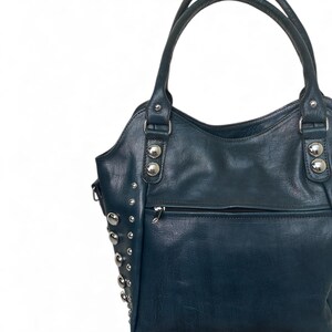 May include: A dark teal handbag with silver-colored studs and hardware. The bag has two handles and a zippered pocket on the front. The leather-like material has a slight sheen.
