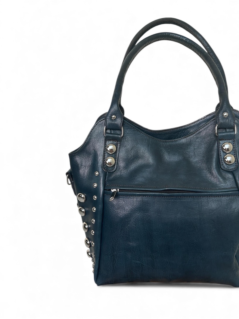 May include: A dark teal handbag with silver-colored studs and hardware. The bag has two handles and a zippered pocket on the front. The leather-like material has a slight sheen.