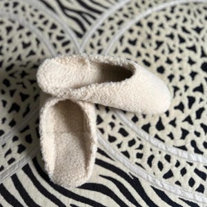 May include: A pair of cream-colored fuzzy slippers. The slippers are made of a soft, plush material and have a simple, rounded shape.