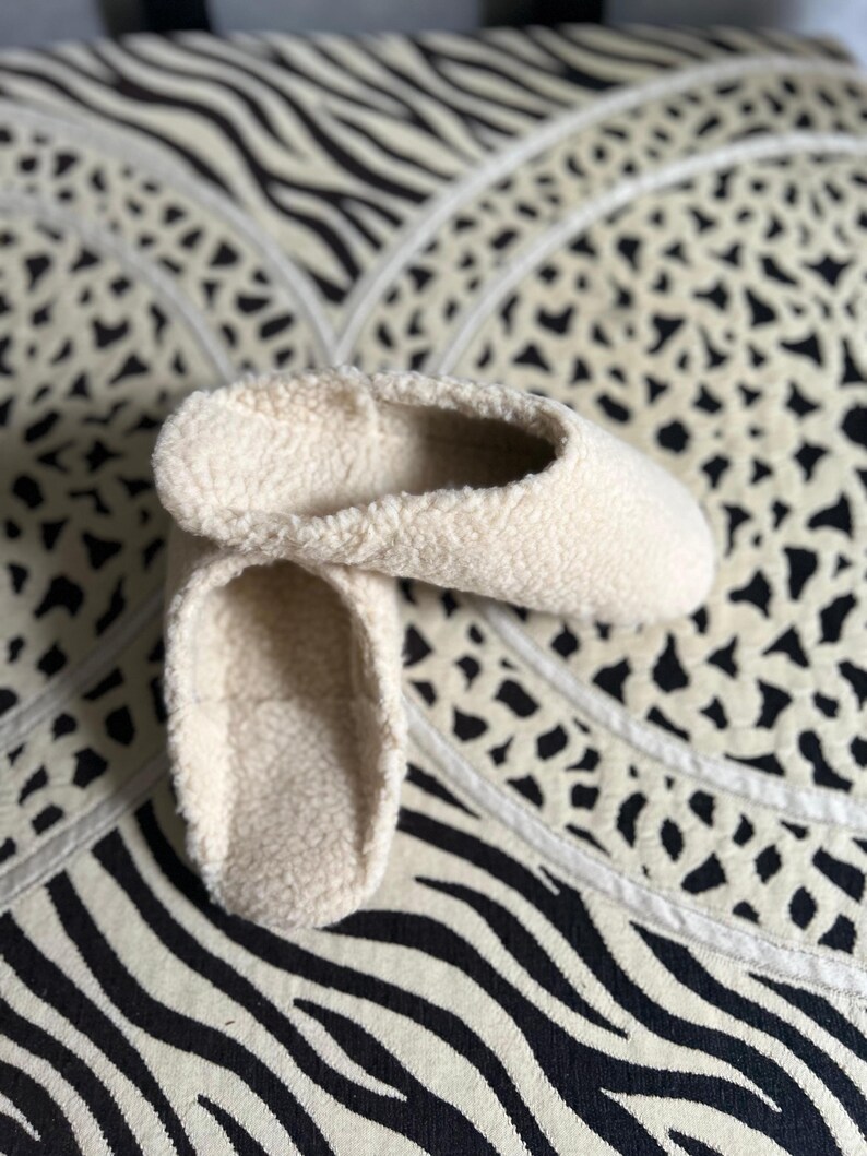 May include: A pair of cream-colored fuzzy slippers. The slippers are made of a soft, plush material and have a simple, rounded shape.