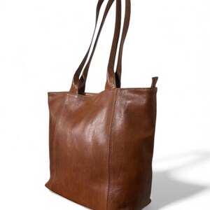 May include: A brown leather tote bag with long, slender handles. The bag has a rectangular shape with a slightly rounded bottom and visible stitching along the seams. The leather has a smooth, slightly distressed appearance.