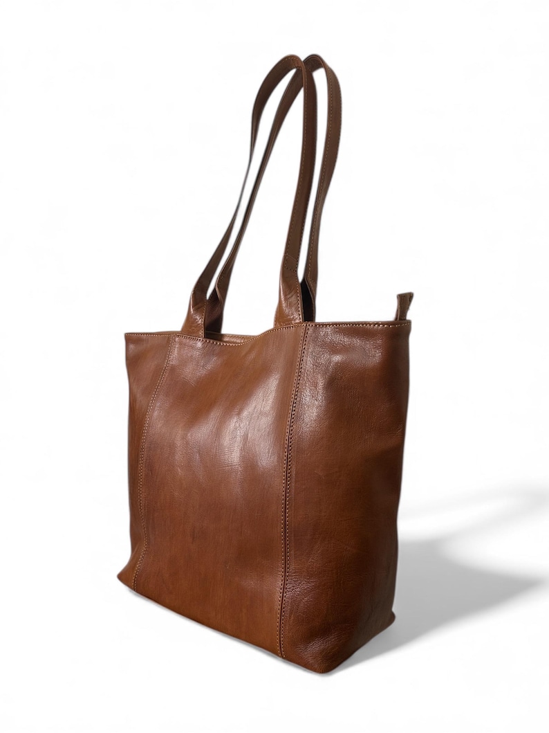 May include: A brown leather tote bag with long, slender handles. The bag has a rectangular shape with a slightly rounded bottom and visible stitching along the seams. The leather has a smooth, slightly distressed appearance.