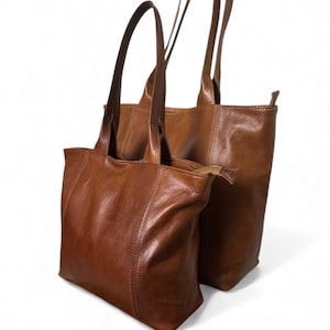 May include: Two brown leather tote bags with long handles. The bags are different sizes, with one slightly larger than the other. The leather has a rich, warm tone and a subtle sheen. The bags are designed for carrying items.