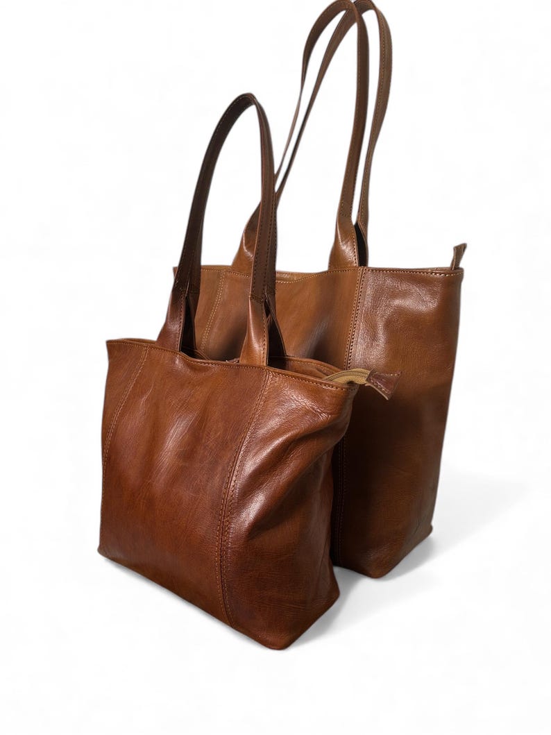 May include: Two brown leather tote bags with long handles. The bags are different sizes, with one slightly larger than the other. The leather has a rich, warm tone and a subtle sheen. The bags are designed for carrying items.