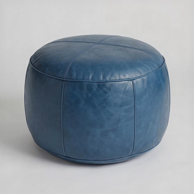 May include: A round, blue leather ottoman. The ottoman has a stitched cross pattern on top and vertical seams. The color is a deep, saturated blue with a slightly distressed appearance. The design is simple and modern.
