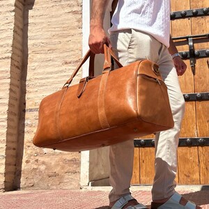 May include: A large, brown leather duffel bag with top handles and a side zipper. The bag is carried by a person wearing light-colored pants and sandals. The background features a brick wall and a wooden door.