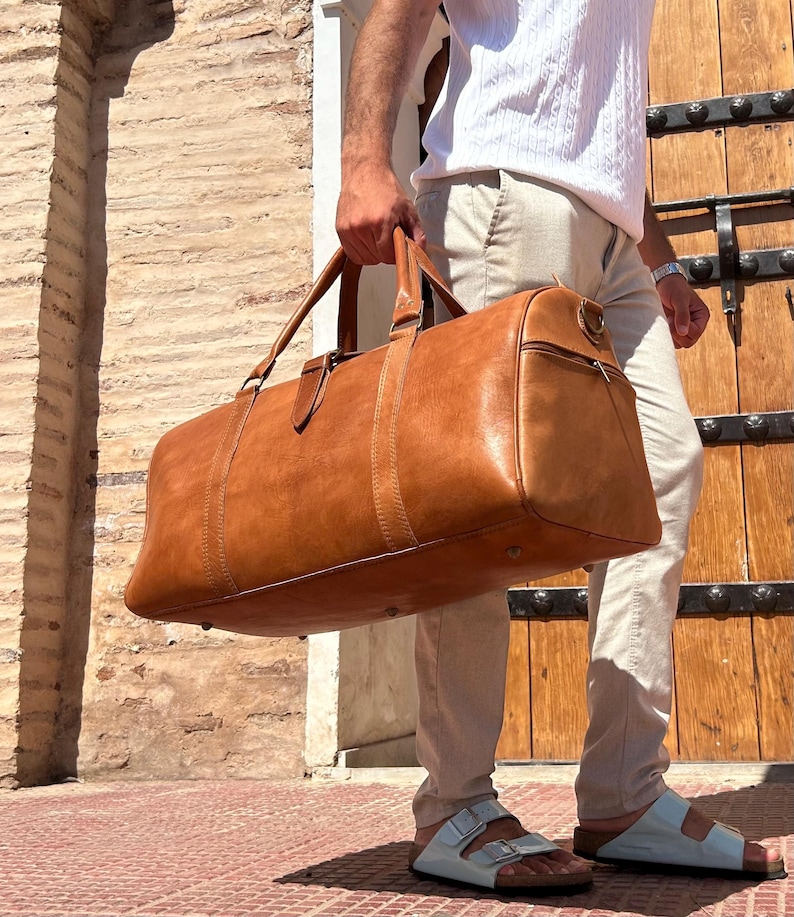 May include: A large, brown leather duffel bag with top handles and a side zipper. The bag is carried by a person wearing light-colored pants and sandals. The background features a brick wall and a wooden door.