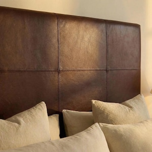 May include: A brown leather headboard with a stitched grid pattern. The headboard is paired with several beige pillows. The headboard is a rectangular shape and is a dark brown color.
