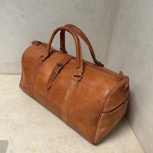 May include: A brown leather duffel bag with two top handles and a shoulder strap. The bag has a cylindrical shape with a zipper closure and a side pocket. The leather has a natural, textured appearance.
