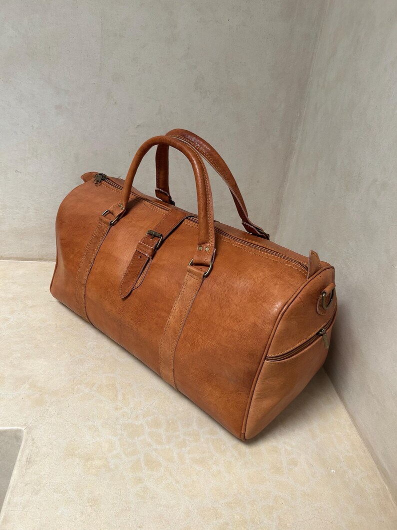 May include: A brown leather duffel bag with two top handles and a shoulder strap. The bag has a cylindrical shape with a zipper closure and a side pocket. The leather has a natural, textured appearance.